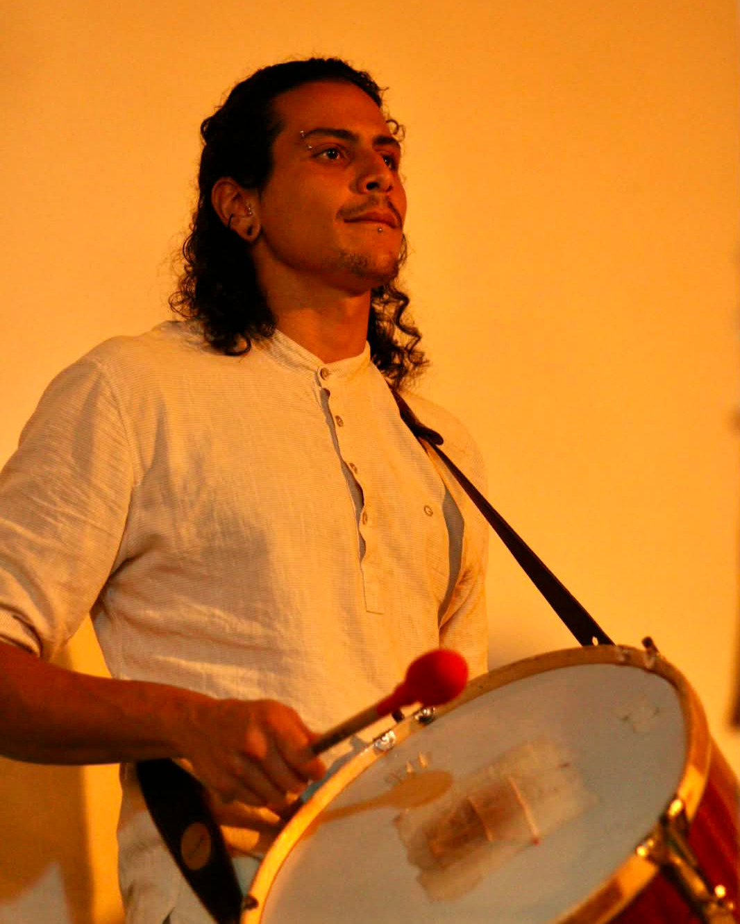 Pedro Petindá em show com a banda Manguacera, usando camisa clara, cabelos cacheados pretos amarrados, pele morena e olhos escuros, com sorriso.